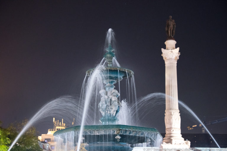 Illuminated Rossio fountain spraying water beside the tall Monument to Pedro IV at night.