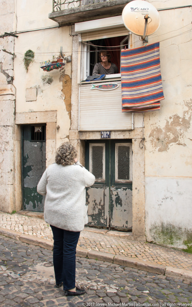 Person leaning from an upper window toward a visitor below on a steep cobbled street with worn façades.