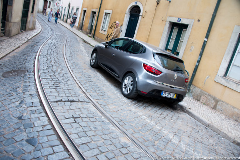Curving tram rails in cobblestones beside a parked gray car and yellow residential façade.