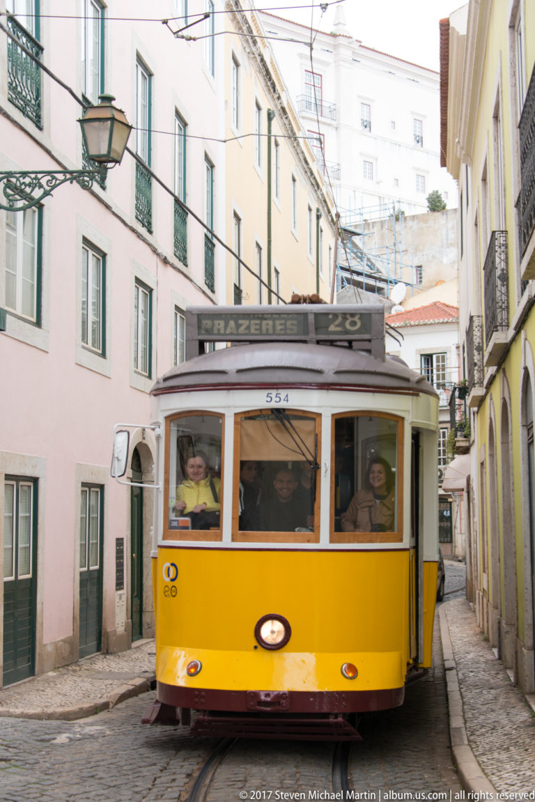 Yellow Lisbon tram approaching on a narrow street with destination board reading 'Prazeres 28.'