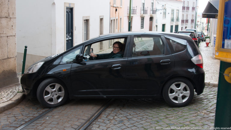 Black compact car turning across tram rails on a narrow cobbled Lisbon street.