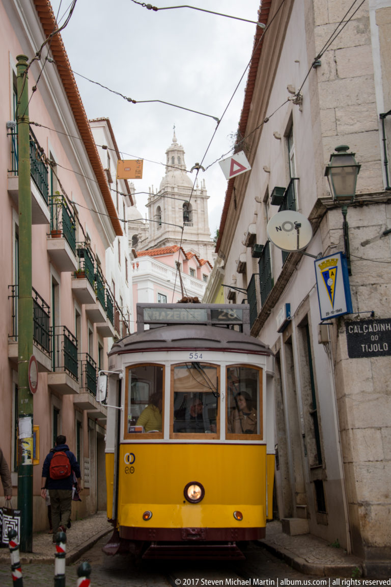 Yellow Lisbon tram approaching on a narrow street beneath wires, with a church façade rising in the distance.