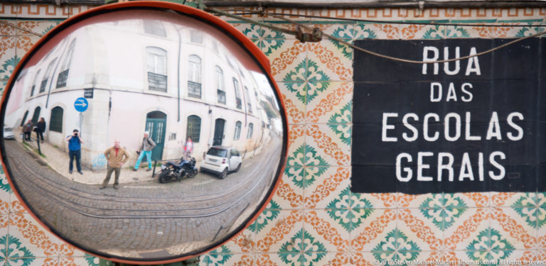 Convex traffic mirror reflecting a Lisbon street beside an azulejo wall and 'Rua das Escolas Gerais' sign.