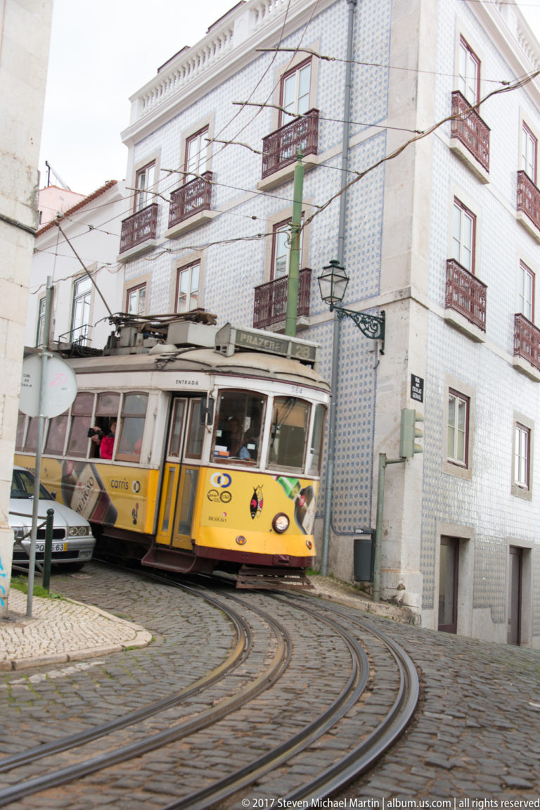 Yellow Lisbon tram curving through rails beside a large blue-and-white tiled corner building.