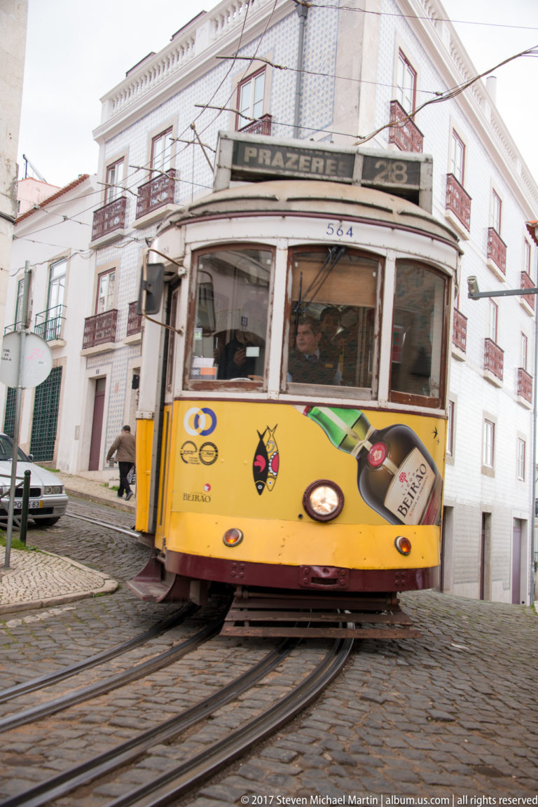 Yellow tram marked 'Prazeres 28' rounding a tight curve on cobbled rails.