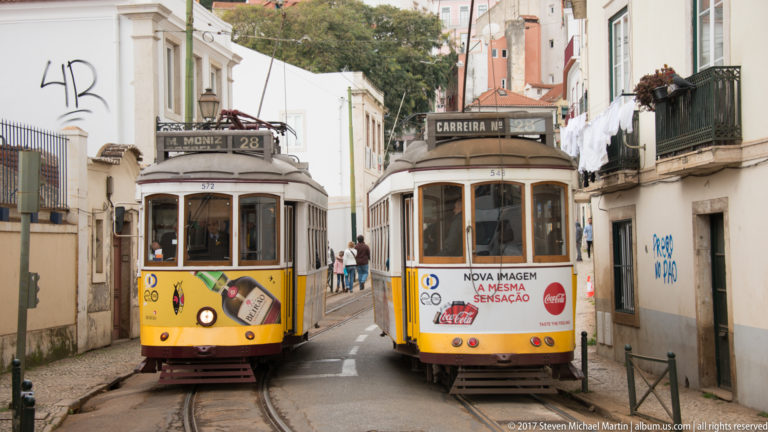Two yellow Lisbon trams passing closely on a narrow street with overhead wires and residential balconies.
