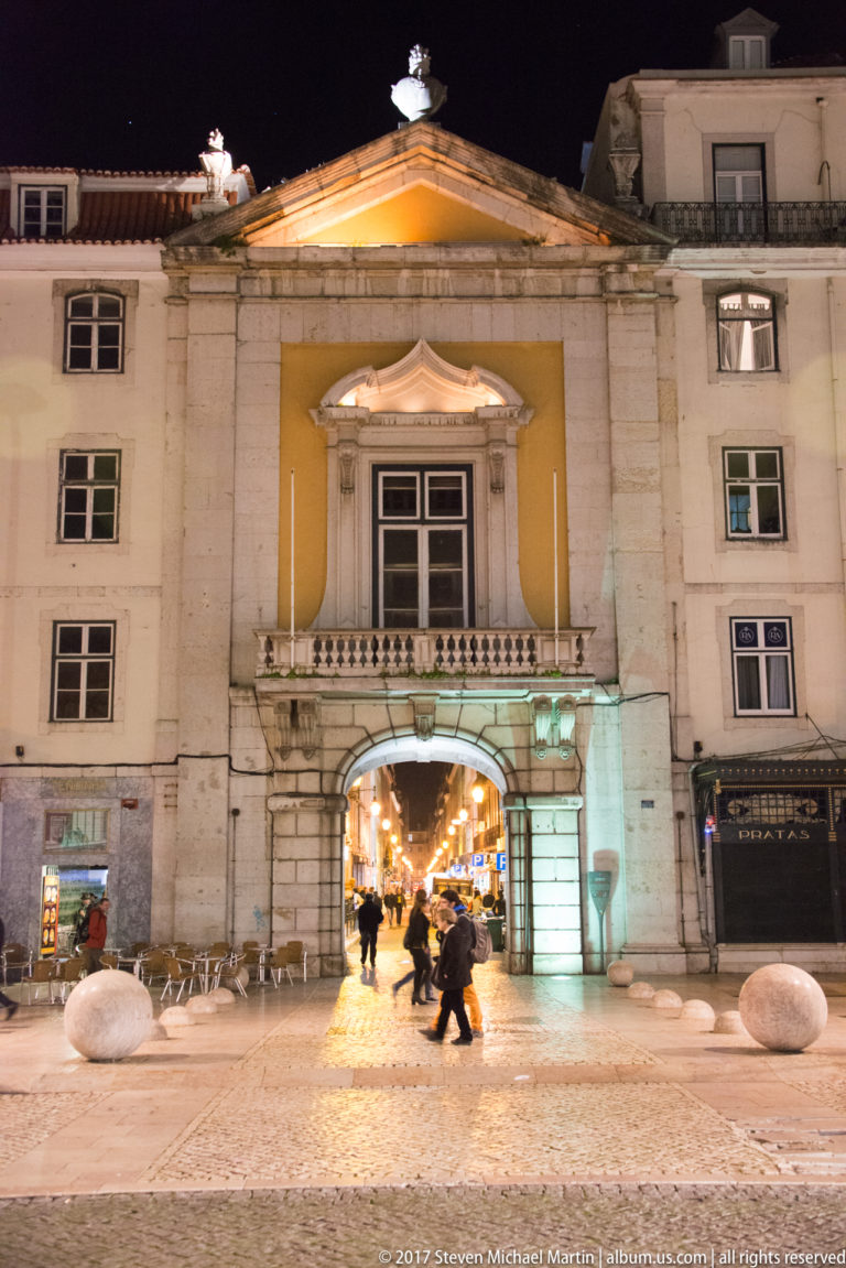 Symmetrical historic façade with a large central archway opening onto a brightly lit street, with pedestrians crossing at night.