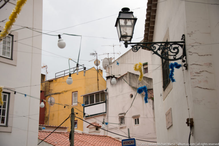 Black decorative wall lantern projecting above white and ochre Lisbon houses with wires, antennas, and hanging decorations.