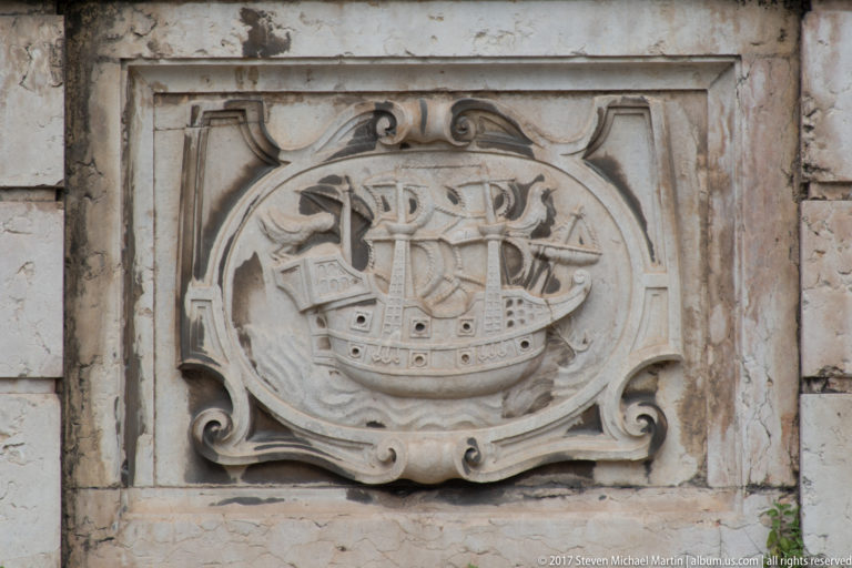 Stone relief of a multi-masted sailing ship inside an ornate scroll-edged cartouche.