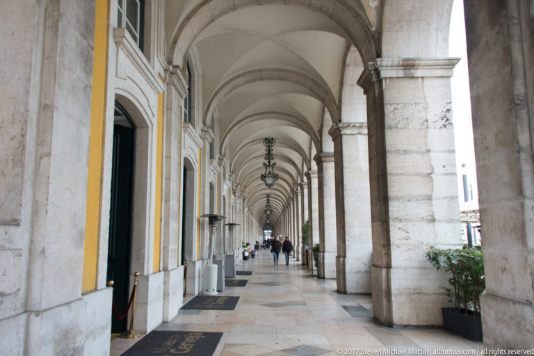 Long stone-vaulted arcade with yellow walls, hanging lamps, doorways, and pedestrians at Praça do Comércio.