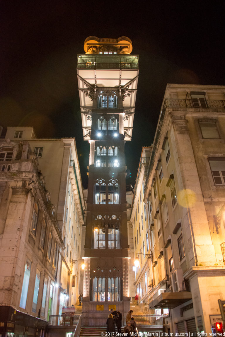 Tall illuminated iron Santa Justa Lift rising between stone buildings on a narrow Lisbon street at night.
