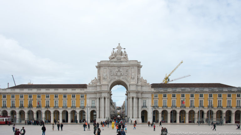 Wide view across Praça do Comércio toward the Arco da Rua Augusta between symmetrical yellow arcaded buildings.