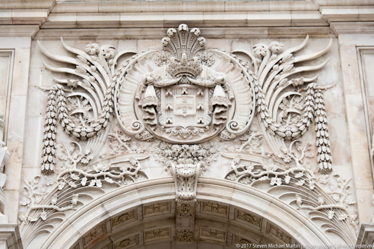 Carved crowned Portuguese royal coat of arms surrounded by foliage and scrollwork above the Rua Augusta Arch.