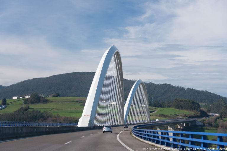 Roadway curves beneath two white steel arch ribs with vertical hangers, with green fields and wooded hills beyond.