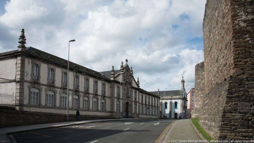 Long pale stone institutional building lines a quiet street opposite the tall rough-stone Roman wall in Lugo.