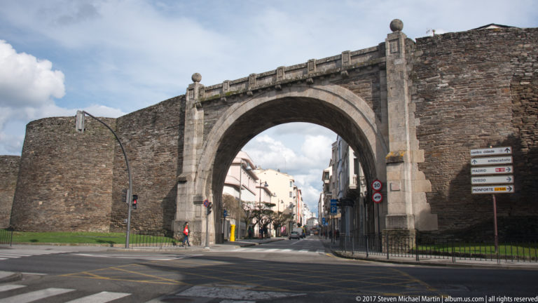 Monumental dressed-stone arch of Porta de Santiago through Lugo’s Roman wall, flanked by rounded towers and carrying a city street.