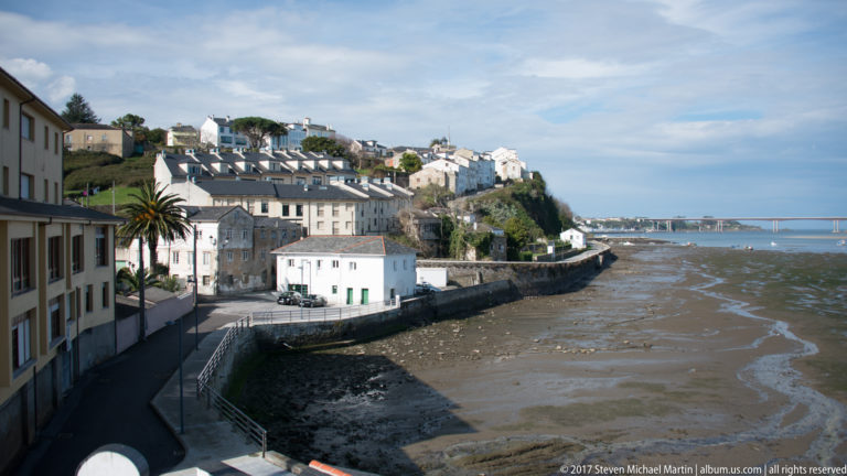 White houses descend to a stone waterfront beside exposed tidal flats, with a palm tree and distant bridge across the estuary.