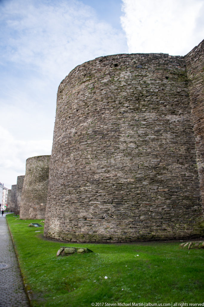 Large rounded Roman wall tower seen close from a grassy perimeter, with additional stone towers receding at left.