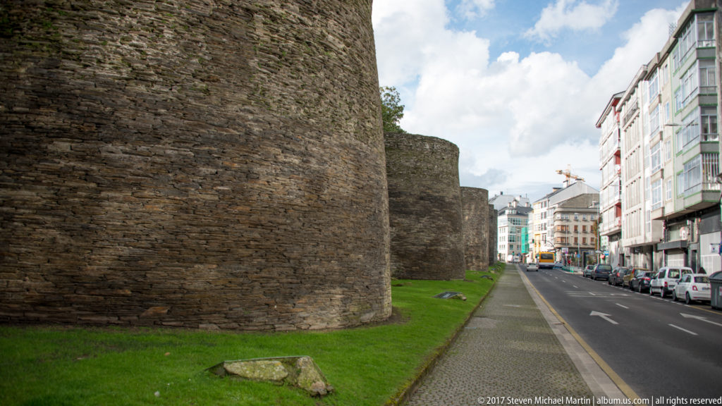 Rounded stone towers of Lugo’s Roman wall line a grass verge beside a paved city street and modern buildings.