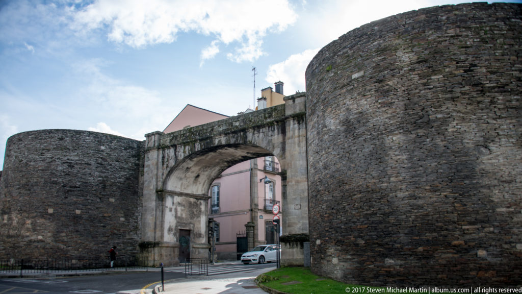 Large arched gateway through Lugo’s stone Roman wall between rounded towers, with a street and buildings visible beyond.