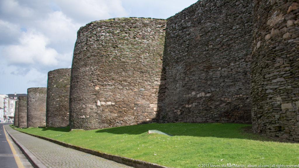 Oblique view along a long row of rounded Roman wall towers beside grass and a narrow street in Lugo.