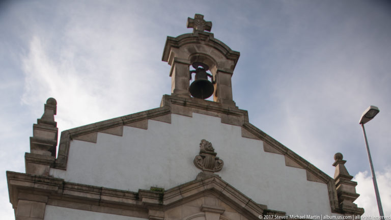 White church gable with granite trim, a single bell in an arched opening, cross, finials, and central relief.