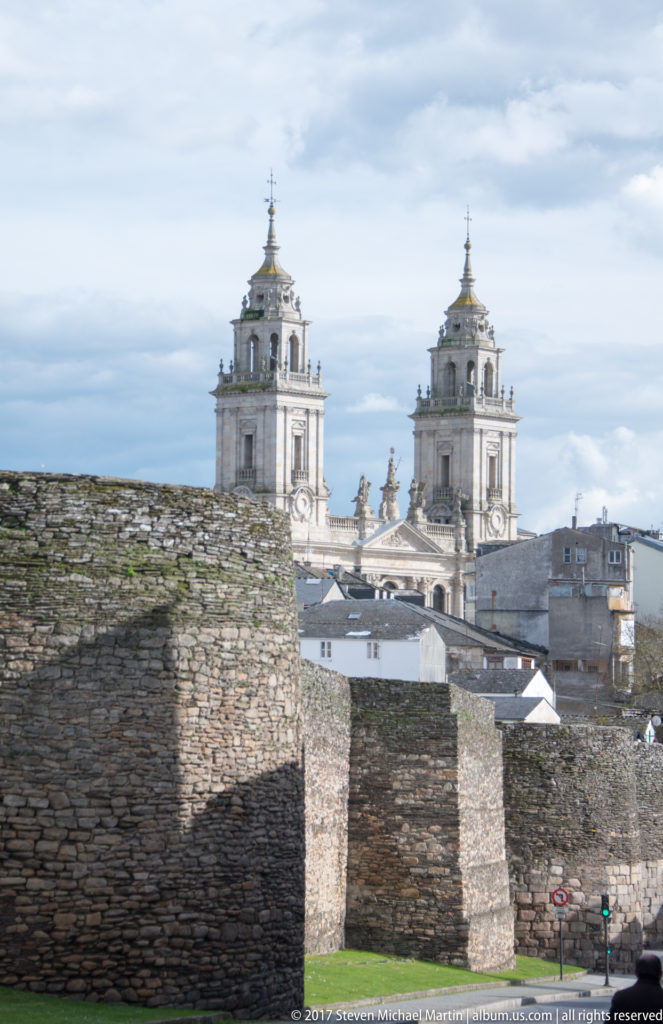 Twin pale stone towers of Lugo Cathedral rise above rooftops and the rough masonry of the Roman wall in the foreground.
