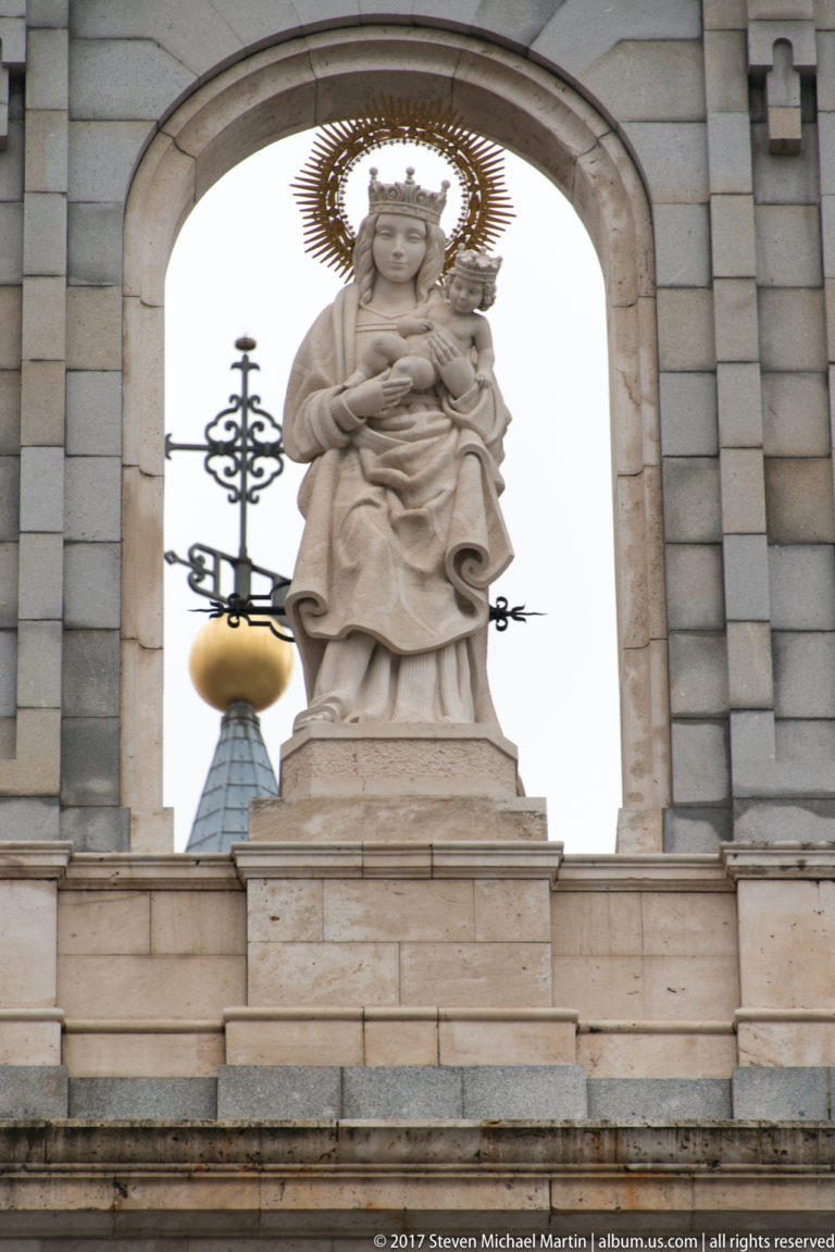 Stone Virgin Mary holding the Christ Child, framed by an arch and radiant metal halo with a decorative cross behind.