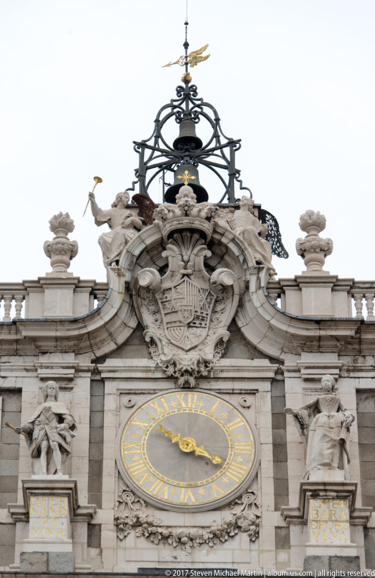 Gilded clock, Spanish royal coat of arms, sculpted figures, and ornamental metalwork crowning the Royal Palace façade.