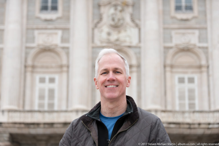 Smiling visitor in a dark jacket standing before the softly blurred stone façade of Madrid’s Royal Palace.