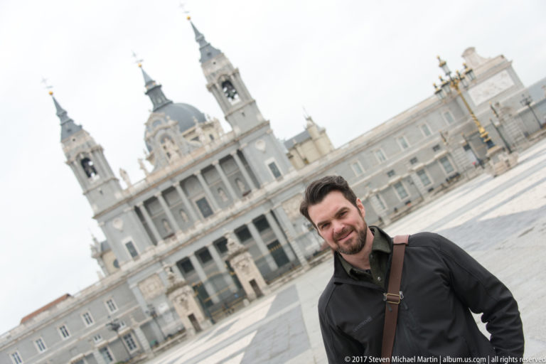 Visitor standing in Plaza de la Armería with the twin towers and dome of Almudena Cathedral behind him.
