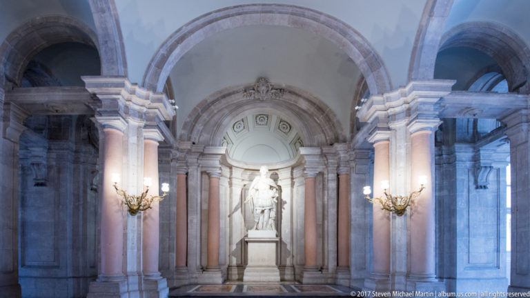 White marble statue on a pedestal inside a large arched niche framed by reddish columns and wall lights.