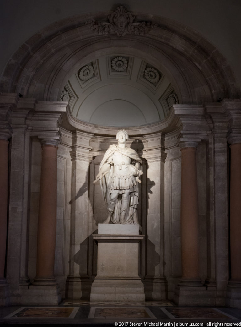 Armored male statue on a pedestal in a dark classical niche, lit strongly from one side.
