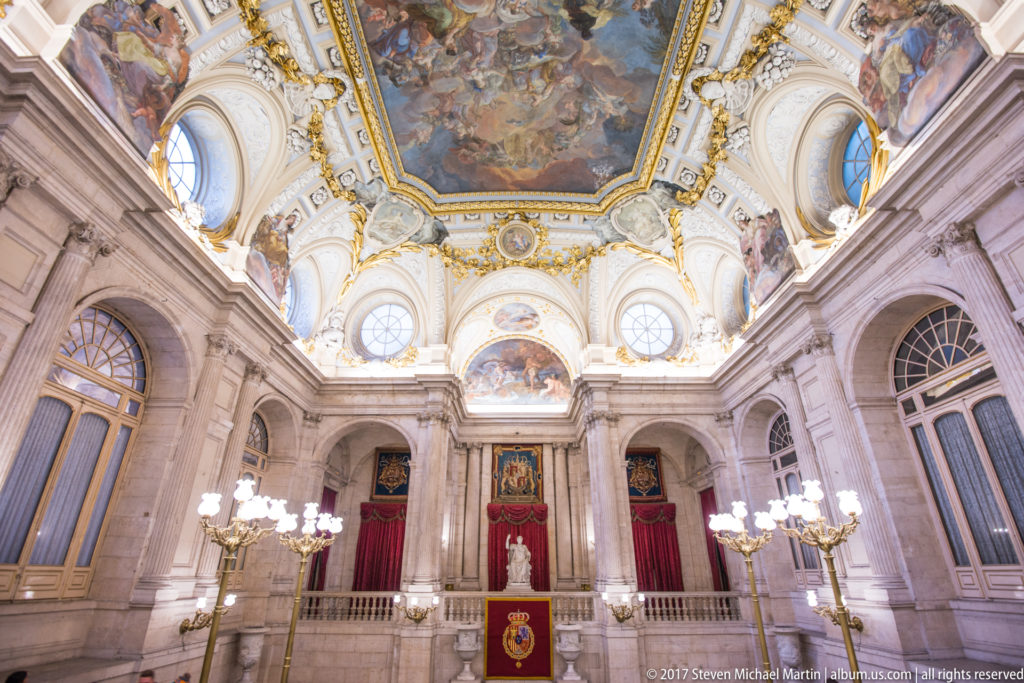 Grand palace hall with ceiling frescoes, gilded ornament, tall windows, chandeliers, red draperies, and heraldic display.