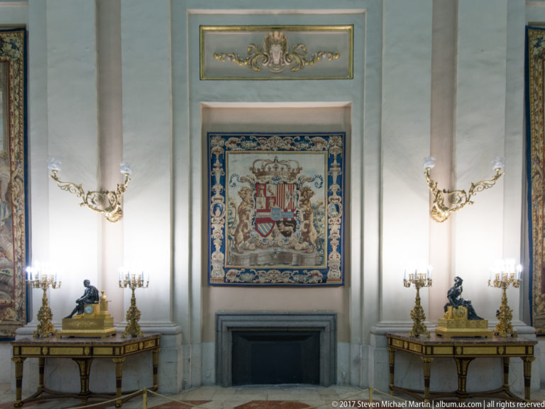 Heraldic tapestry over a dark fireplace, flanked symmetrically by gilt tables, candelabra, sconces, and bronze figures.