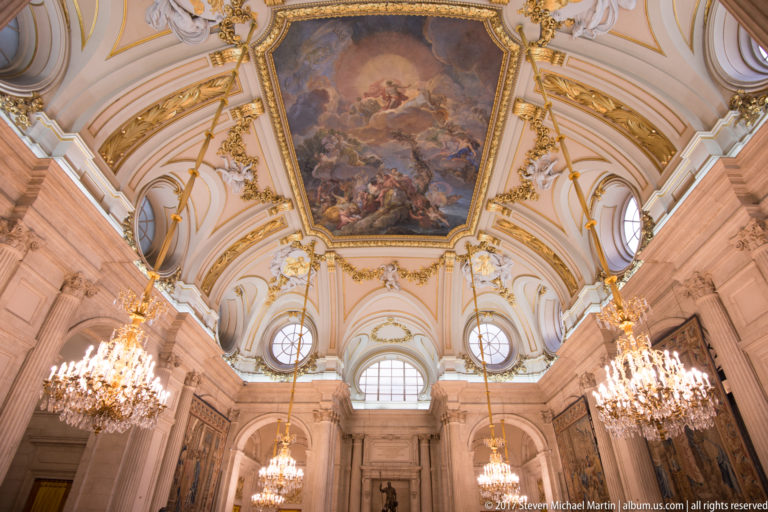 Upward view of a palace ceiling with large fresco, gilded moldings, round windows, and hanging crystal chandeliers.