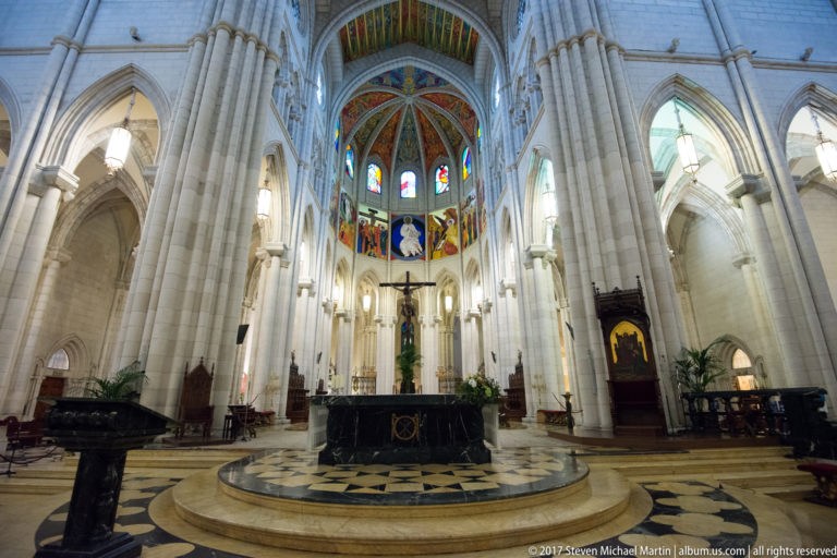 Wide interior view down Almudena Cathedral toward the raised sanctuary, crucifix, stained glass, and colorful apse ceiling.