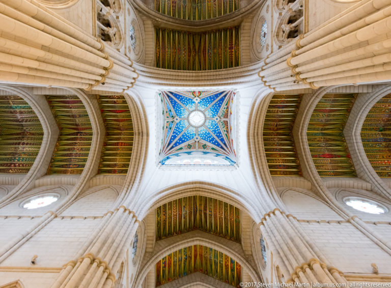 Upward view of Almudena Cathedral crossing with pale stone ribs, blue star-patterned vault, and multicolored ceiling panels.