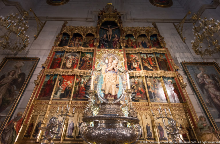 Crowned Marian statue before a gilded multi-panel altarpiece, with paintings and silver-toned liturgical furnishings.