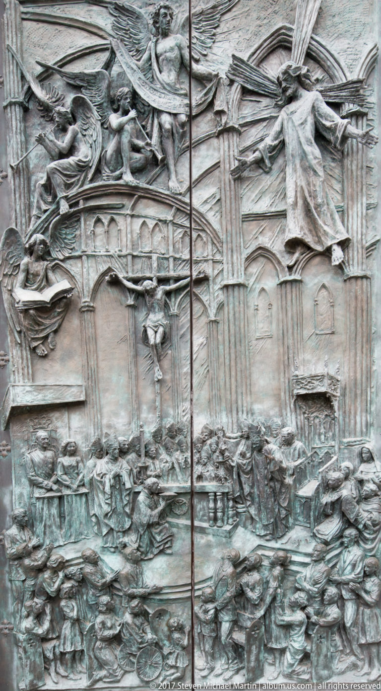 Large bronze-toned relief showing crowds, architecture, and sweeping figures across two joined metal panels.