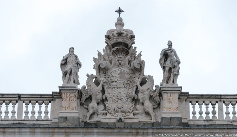 Large carved royal coat of arms on the Royal Palace roofline, flanked by standing statues and ornate stone decoration.