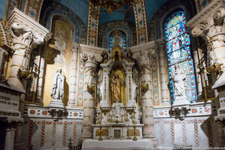 Decorated cathedral side chapel with central Marian altar, white statues, patterned walls, gilded details, and stained glass.