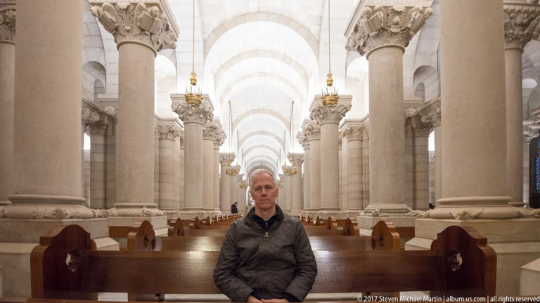Seated visitor centered between rows of pews and massive pale stone columns in a long cathedral nave.