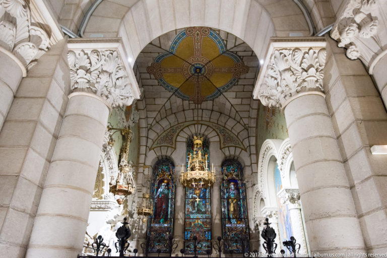 Pale stone columns with carved capitals framing stained glass, a circular patterned window, and a distant devotional shrine.