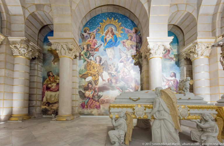 Colorful religious mural behind a white stone tomb or memorial supported by kneeling angel figures between massive columns.