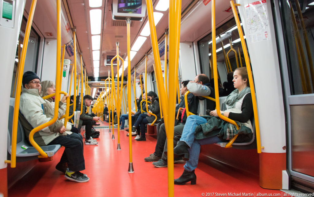 Passengers seated inside a Madrid Metro car with yellow grab poles, facing benches, and a bright red floor.