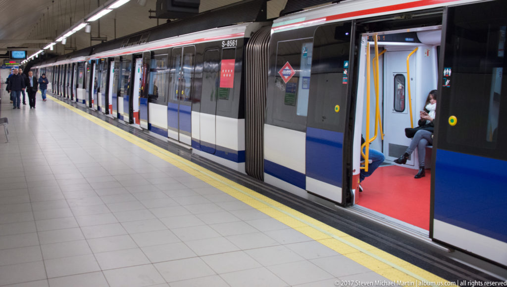 Blue-and-white Madrid Metro train with open doors beside a tiled underground platform and yellow safety line.