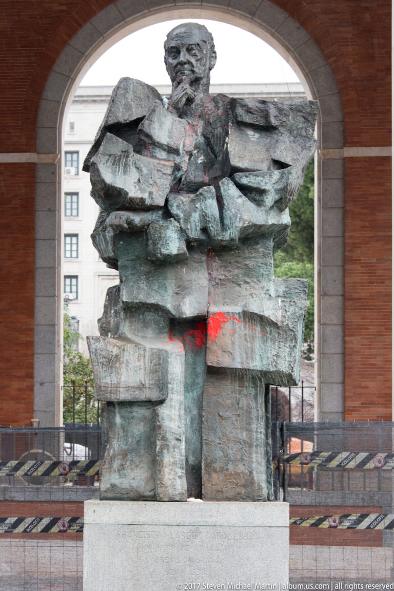 Large blocky bronze human figure on a stone pedestal beneath an architectural arch, with city buildings visible behind.