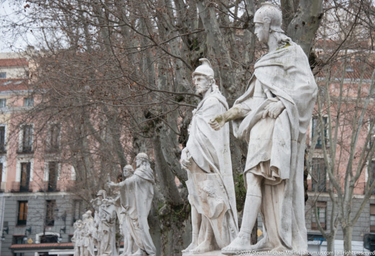 Row of pale stone statues of Spanish monarchs on pedestals among leafless trees in Plaza de Oriente.