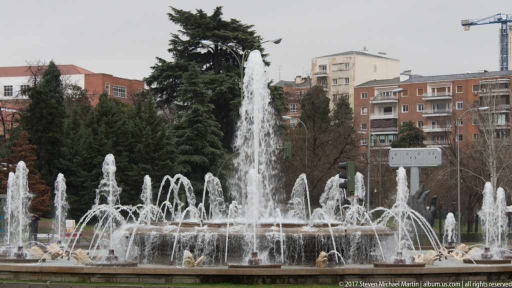Large urban fountain with tall central jet and rings of smaller sprays before trees and apartment buildings.